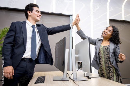 Young businesspeople make high five hand touch over desktop computer in office. Two business staff colleagues greet with hi five. Businessman and businesswoman do high five to celebrate the success.