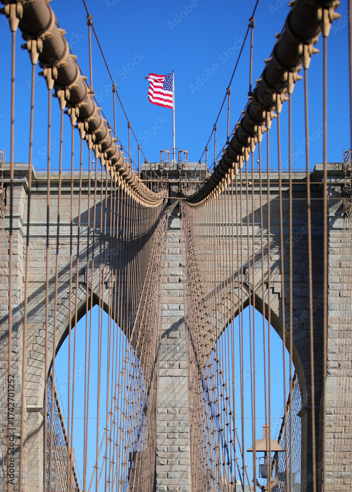 Obraz premium American flag waving above Brooklyn Bridge tower pillar with blue sky and steel cables