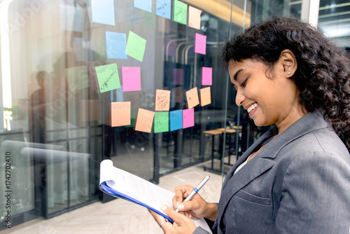 Young businesswoman with curly hair writing her idea on sticky note paper, critical thinking in project work. Beautiful business staff worker working in office. Female employee at workplace.