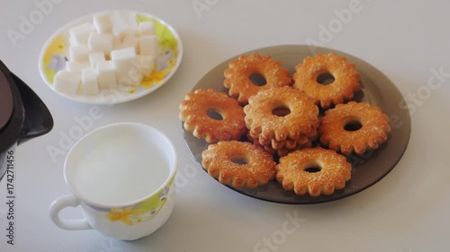 Water is poured from a transparent electric kettle into a white cup on a table with sweets. Medium shot.