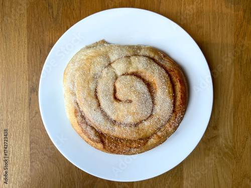 Overhead view of a Norwegian cinnamon roll called Skillingsbolle on a white plate over wooden table, traditional Scandinavian pastry sprinkled with sugar