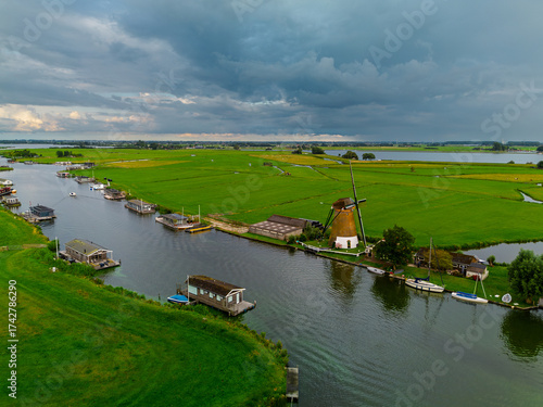 Aerial view of Dutch countryside with traditional windmill, canal, docked boats, and green fields capturing rural charm and water management under dramatic overcast skies.