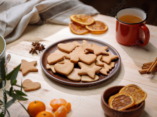Wooden platter with baked gingerbread shapes is surrounded by seasonal spices, oranges, and a mug of tea.