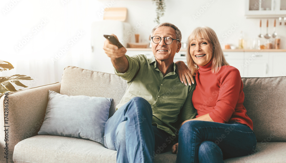 Obraz premium Happy Senior Spouses Watching TV Pointing Television Remote Control Sitting And Hugging On Sofa At Home. Mature Couple Enjoying Favorite Movie Together On Weekend. Retired Family Leisure
