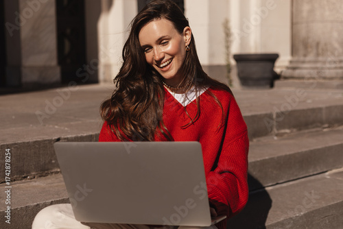 Happy lifestyles young woman freelance smile using laptop computer with smile while working sitting on stairs outdoors in the city. Remote working concept outside the office. Girl wear red sweater.