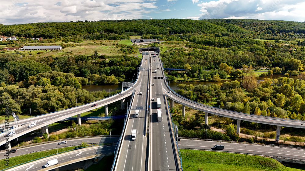 Αυτοκόλλητο Aerial view of highway interchange with elevated ramps and cars, green countryside hills and dramatic sky, modern transport infrastructure network for logistics and mobility concept