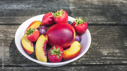 Colorful bowl of fresh fruits including strawberries, plums, and peaches on a wooden table