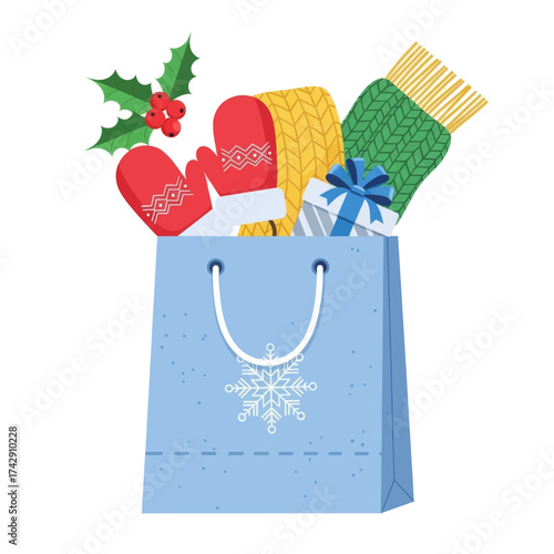 A blue holiday shopping bag with a snowflake, filled with warm winter scarves, mittens, a gift box, and a sprig of holly.