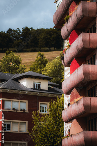 Vertical telephoto view of red and concrete curved balconies with plants, contrasted against nearby residential buildings and green hills, combining urban architecture with nature