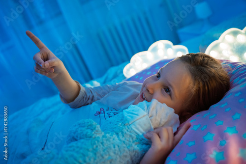 Cute little toddler girl laying in bed with her teddy bear and point at ceiling looking at the stars 