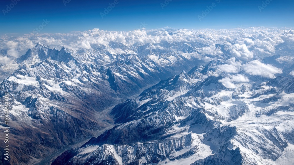 Fototapeta premium Panoramic aerial view of rugged snow covered mountain range with dramatic clouds and blue sky, evoking awe and tranquility