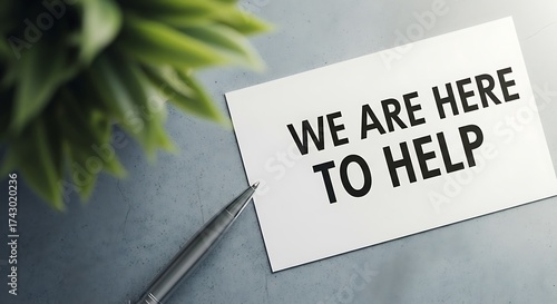 A white card with the text we are here to help written in black, placed on a grey surface next to a pen and a green plant