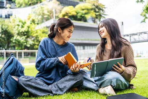 Fotografija Two young asian female college friends are sitting on the grass at university, happily tutoring each other