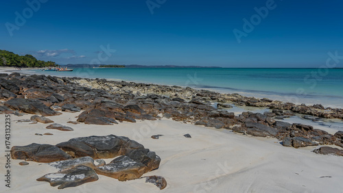 Boulders are scattered on the sandy beach. A calm aquamarine ocean. Motorboats are moored in the distance. Silhouettes of people. Green vegetation, palm trees on the coast. The blue sky. Madagascar.