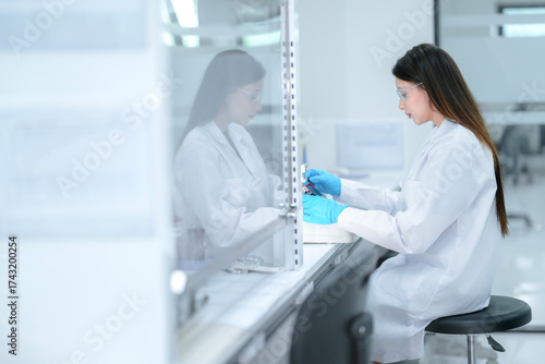 A pharmaceutical technician performs aseptic compounding inside a sterile biosafety cabinet, preparing a cell culture for biologic drug development under strict quality control conditions.