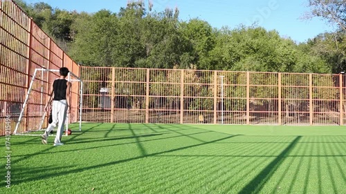Close action as one player blocks the other attempt to advance red ball near goal. Both wear black, move swiftly over green turf under bright sun.