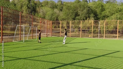 Opposing players confront each other in energetic contest for red ball near goal. Summer sun, urban cage field, green synthetic grass, black sportswear.