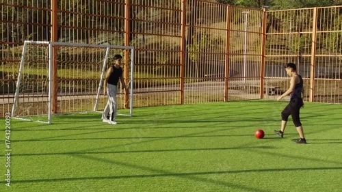 Two men on football pitch prepare for penalty. One lines up shot, goalkeeper observes with alert stance. Bright sunlight, sportswear, orange fence, red ball. Scene of strategy and rivalry.