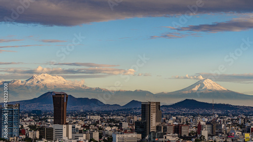 A city with mountains in the background. The sky is blue and the clouds are white. Panoramic view of Mexico City, snow-covered volcanoes Popocatepetl and Iztaccihuatl