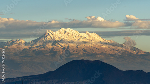 A mountain range with snow on the top and a cloudy sky. The mountains are in the distance and the sky is blue. Panoramic view of Mexico City, snow-covered volcanoes Popocatepetl and Iztaccihuatl