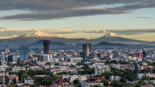 A city with a mountain range in the background. The city is full of tall buildings and the sky is cloudy. Panoramic view of Mexico City, snow-covered volcanoes Popocatepetl and Iztaccihuatl