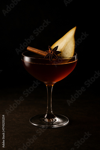A signature Thanksgiving cocktail in an elegant coupe glass. The drink is a deep amber color, garnished with a thin slice of pear, a cinnamon stick, and a star anise.