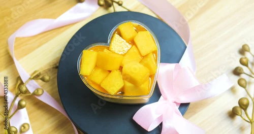 Heart shaped mango dessert sprinkled with golden accents, tied with pink ribbon, resting on dark plate against wooden background, symbolizing romantic sweetness