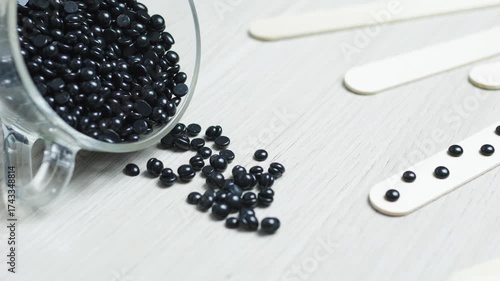Black hard wax beans are spilling from a transparent glass jar onto a light wooden table, with wooden spatulas nearby, ready for a depilation session