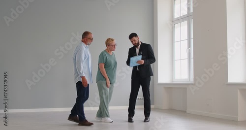 Realtor holds consultation with senior couple of buyers during a tour and viewing of an empty apartment interior, noting layout. Real estate agent showing house to elderly clients.