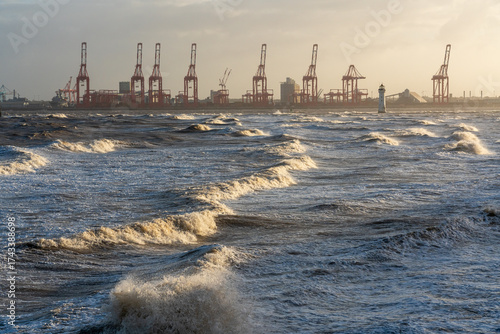 Rough sea waves from Storm Amy crashing in front of the Port of Liverpool with container cranes and Perch Rock lighthouse visible in golden morning light