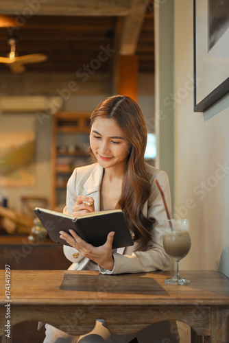Smiling businesswoman in beige blazer sitting at a wooden table, taking notes in a notebook