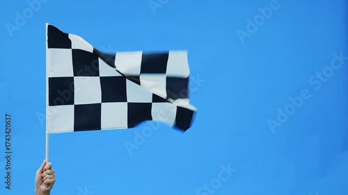 Human hand waving checkered flag on blue sky background, slow motion video