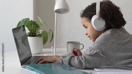 Primary school African American Student child with headphones hand writing in book using laptop. Distance learning online education. School girl studying at home with digital tablet computer and doing