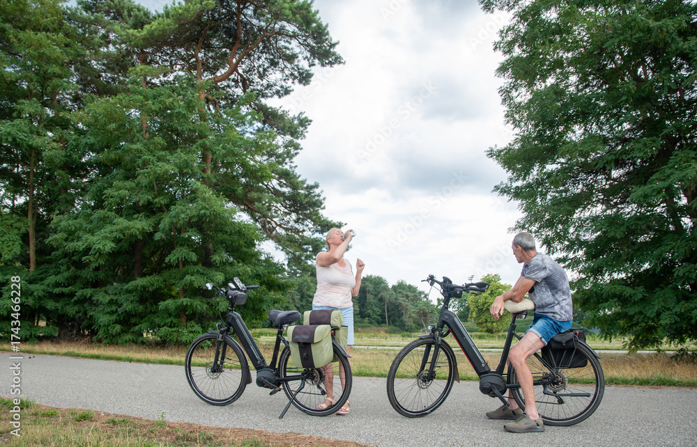 Obraz premium Elderly couple enjoys active retirement by riding modern electric bikes along scenic nature trails, while a woman drinks water and relaxes in the fresh air during summer vacation