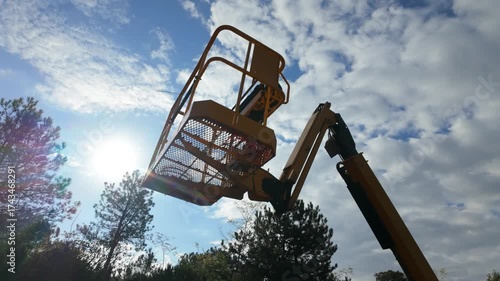On a sunny day, a manlift platform extends upward toward the sky, with clouds and trees in the background adding depth and atmosphere to the scene.