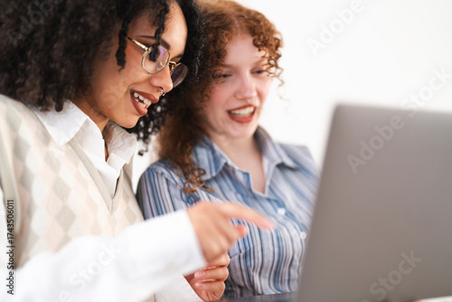Two young women collaborate on a laptop, one pointing at the screen with a smile, sharing ideas and working together on a project.