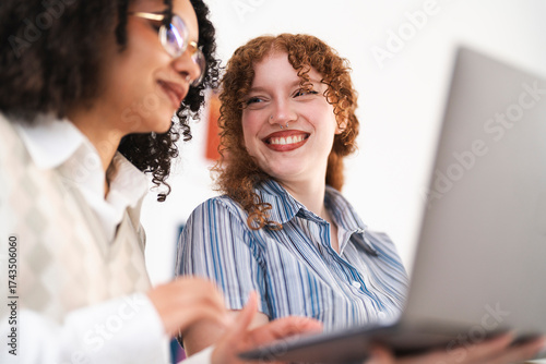 Two young women with curly hair collaborate on a laptop, sharing ideas and smiling in a bright, modern setting.