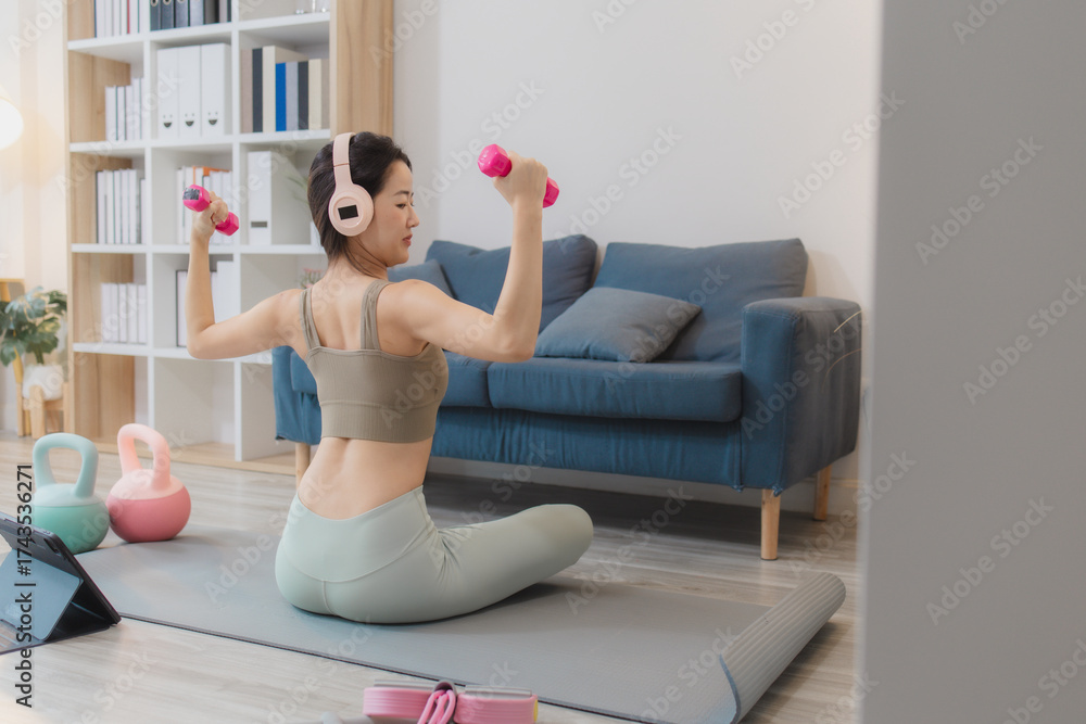 Fototapeta premium A young woman is exercising at home, practicing yoga and strength training with dumbbells and kettlebells on a mat. She follows her workout routine with focus and determination.