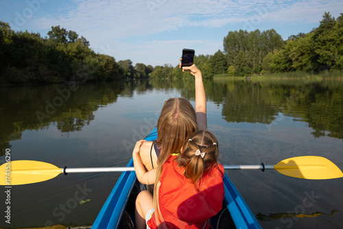 Mother taking a selfie with her daughter while kayaking on a calm lake surrounded by green trees on a sunny summer day. Happy family outdoor recreation and nature adventure