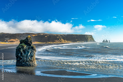Basalt sea stack rises from the black sands of Reynisfjara Beach near Vík í Mýrdal, Iceland, with Atlantic waves and Reynisdrangar sea stacks in the distance.