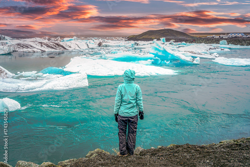 A traveler in a turquoise jacket watches floating icebergs at Jokulsarlon glacier lagoon in Iceland during a colorful sunset, symbolizing adventure and solitude.