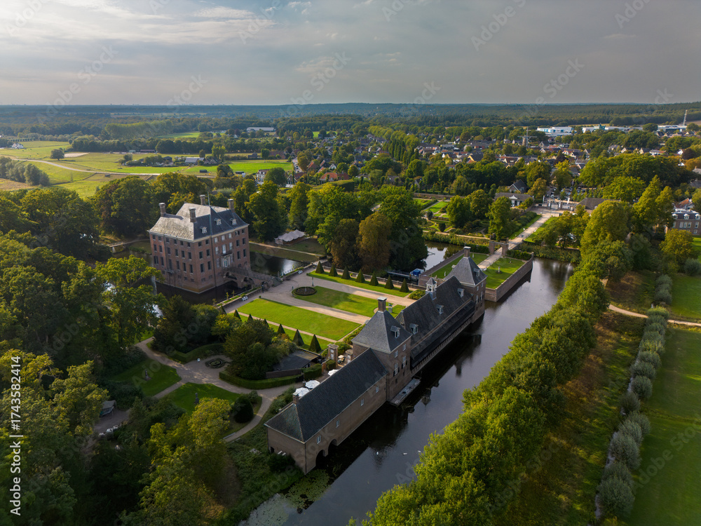 Fototapeta premium Aerial view of historic moated castle with gardens, bridge, and adjacent long building, surrounded by trees, water features, and a nearby village under warm sunlight.
