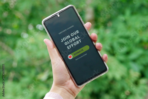 Hand holding smartphone displaying charity message build a brighter future donate button with green nature background sunlight