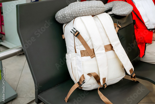 White travel backpack with brown straps and gray neck pillow on airport waiting area seat, travel essentials concept
