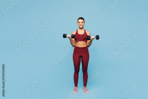 Fotografie Full body young fitness trainer instructor sporty woman sportsman wears red sportswear training in home gym hold dumbbells lift weight isolated on plain blue background