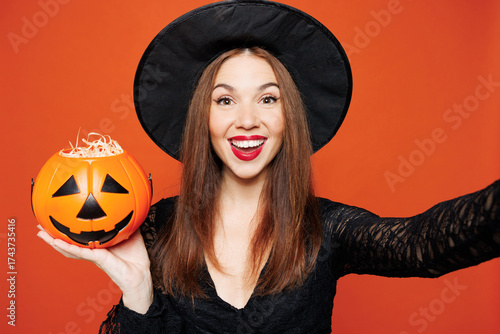 Close up young woman wears black hat dress witch costume doing selfie shot pov on mobile cell phone hold pumpkin isolated on plain red orange background. Celebration Halloween holiday party concept.