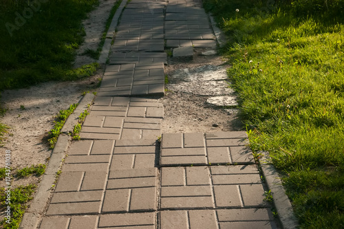 Broken tiled path leading into the distance with grass on both sides, parallel dirt trail on the left, deep shadows and a bright sunlit patch across the path.