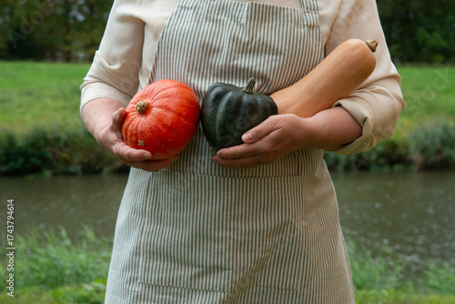  Autumn harvest. A woman in an apron holds in her hands three pumpkins of different shapes and colors against the background of nature,