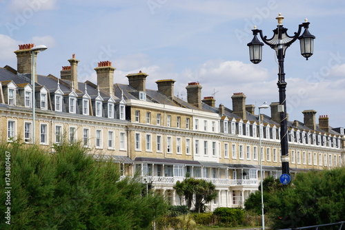 A row of traditional terraced houses with bay windows and balconies, overlooking a green area with lush foliage and a lamppost in Worthing in the UK England