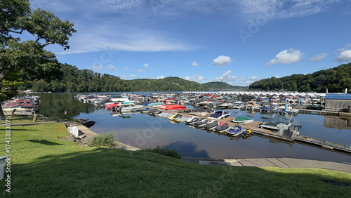A wide, eye-level shot of a marina on a bright, sunny day. The calm, reflective water is filled with rows of docked boats, including pontoon boats, sailboats, and a few jet skis. 
